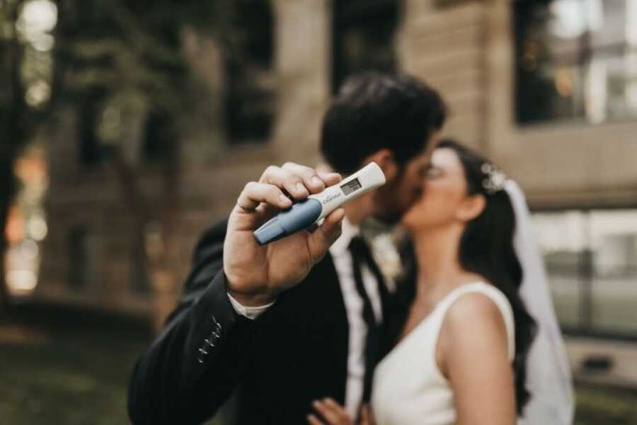 couple stands together on their wedding day holding up a pregnancy test