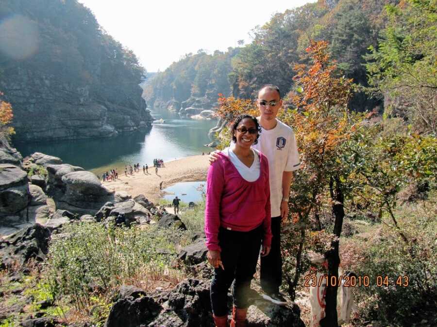 A visually impaired man stands with his wife outdoors near a river