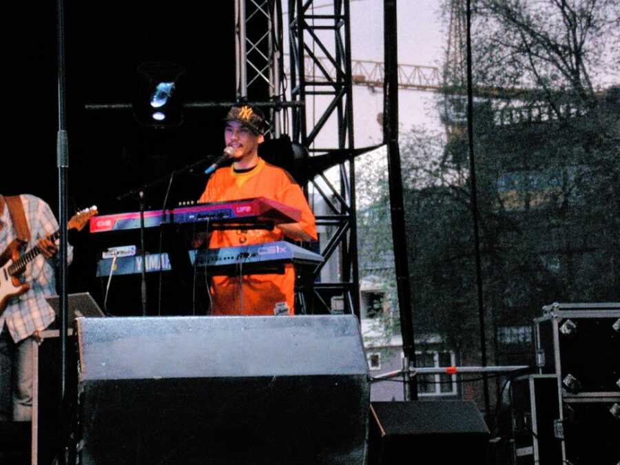 A man on stage wearing an orange shirt and Yankees cap playing the keyboard 