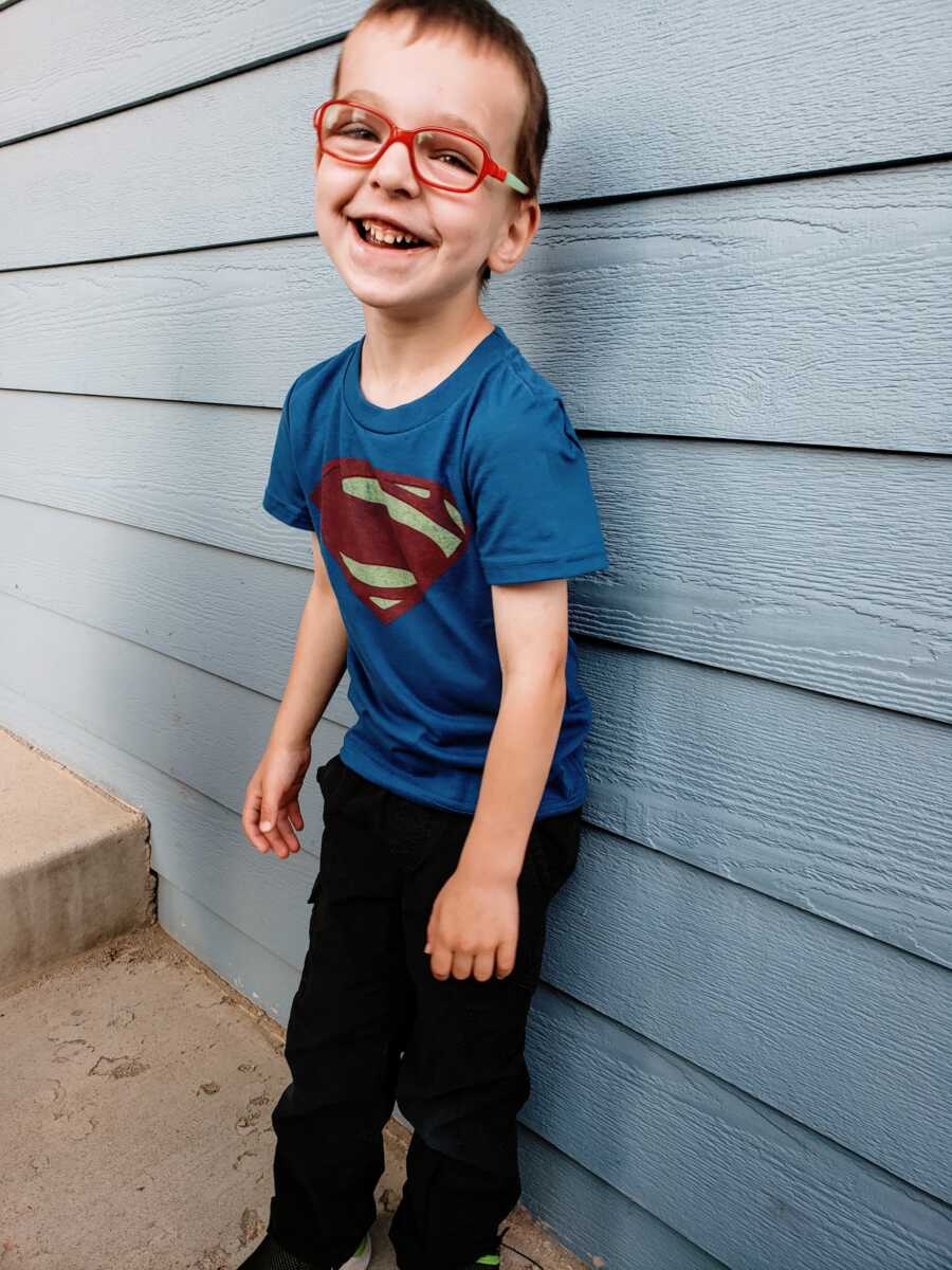A boy wearing Superman shirt leans against a house with blue siding