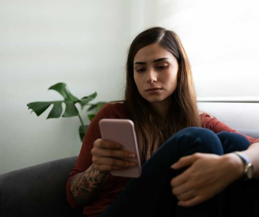 Young woman sits on couch with arm around leg against her chest while she looks at her phone. 