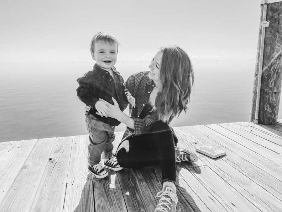 A mom kneels next to her young son who stands on a dock