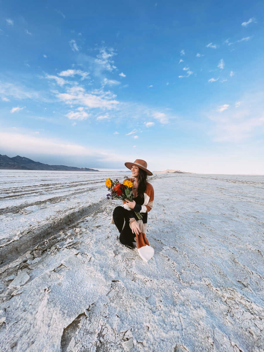 Woman wearing a hat holds flowers and takes picture in the sand.