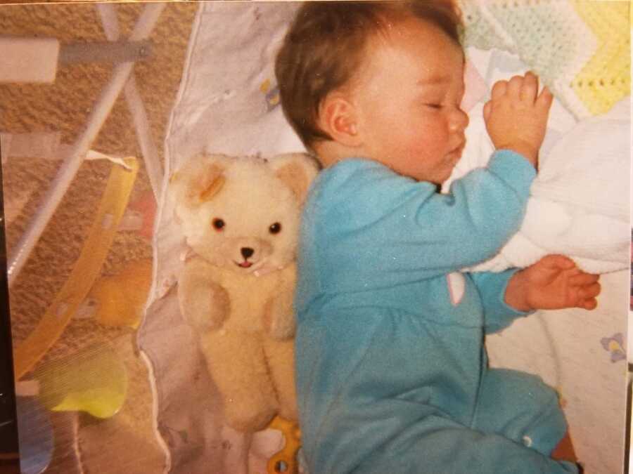 Baby foster girl in blue onesie sleeps next to teddy bear.