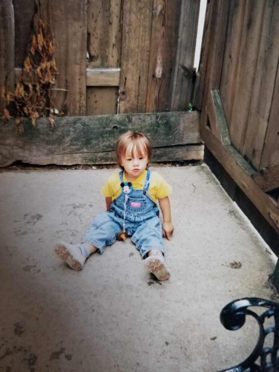 Toddler foster girl in overalls and yellow shirt sits on cement.