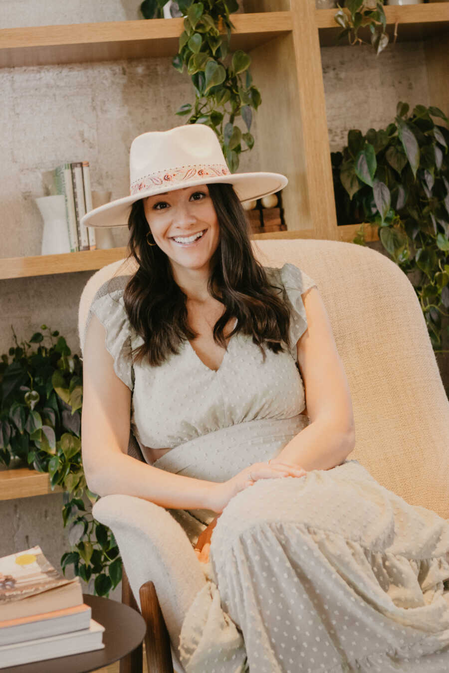 Woman wearing grey dress and hat sits in chair in front of bookshelf.