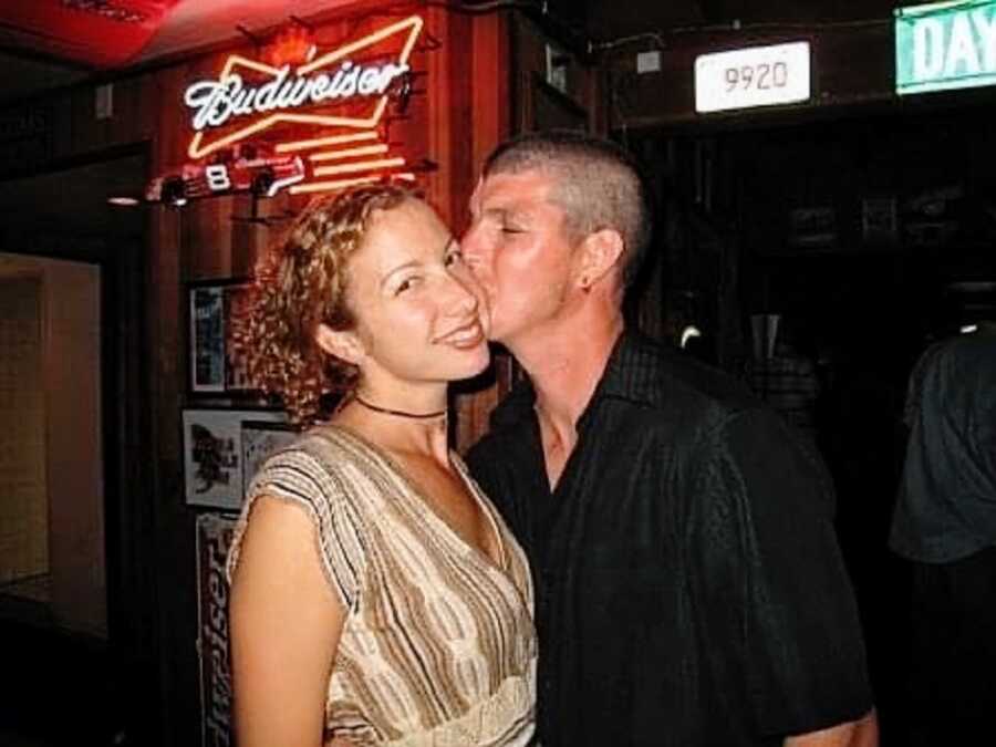 A man kisses his partner on the cheek while both are in a bar