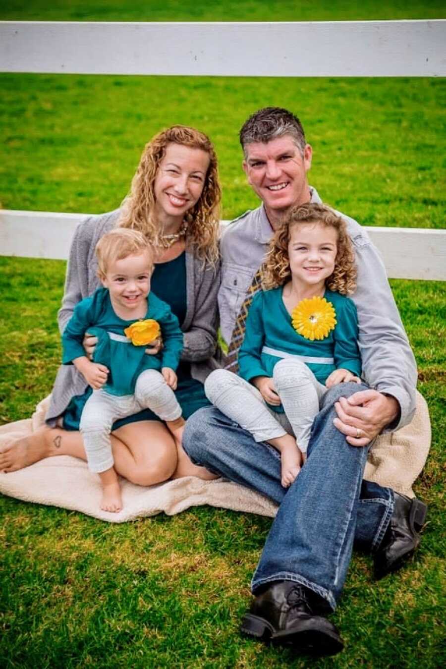 Parents sit by a fence with their two young daughters