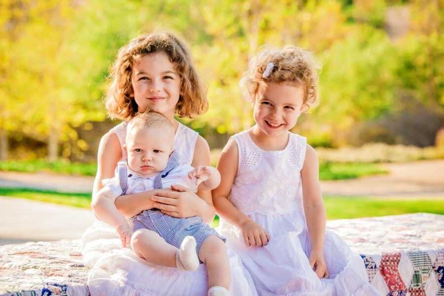 Three young siblings sit together on a blanket outdoors