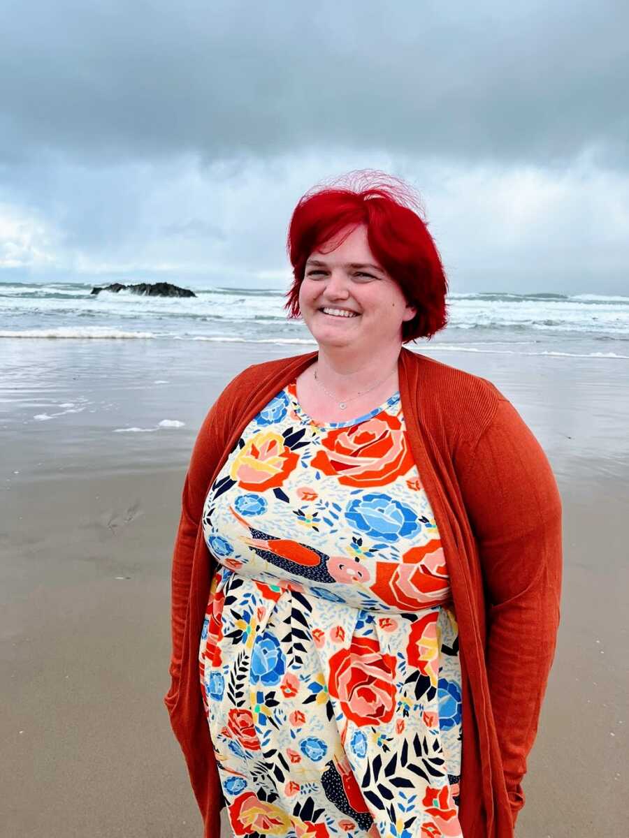 A woman with red hair and a red shirt stands on a beach