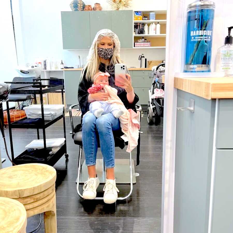 new mom sits at hair salon with newborn on her lap