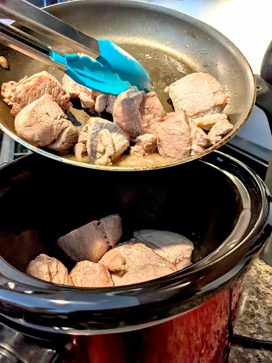 adding the browned pork butt into the slow cooker with tongs