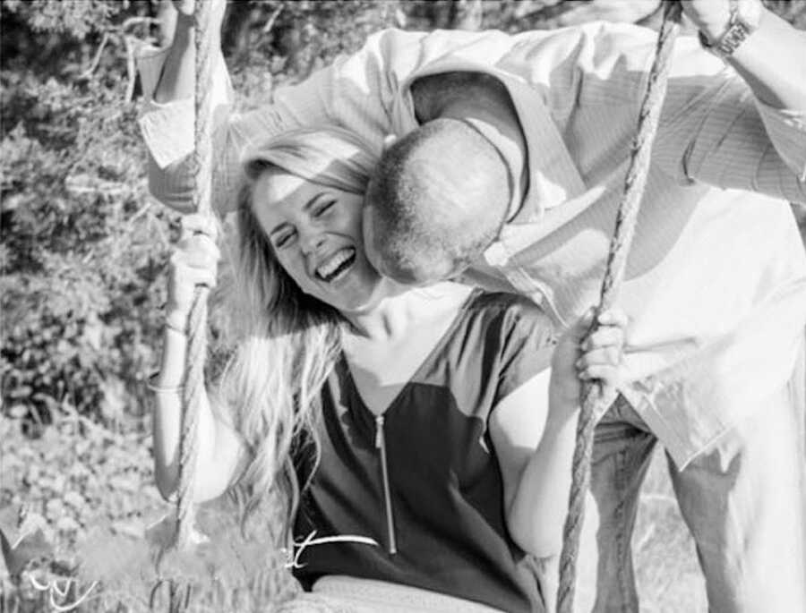 woman sits on swing laughing while husband stands behind and kisses her neck