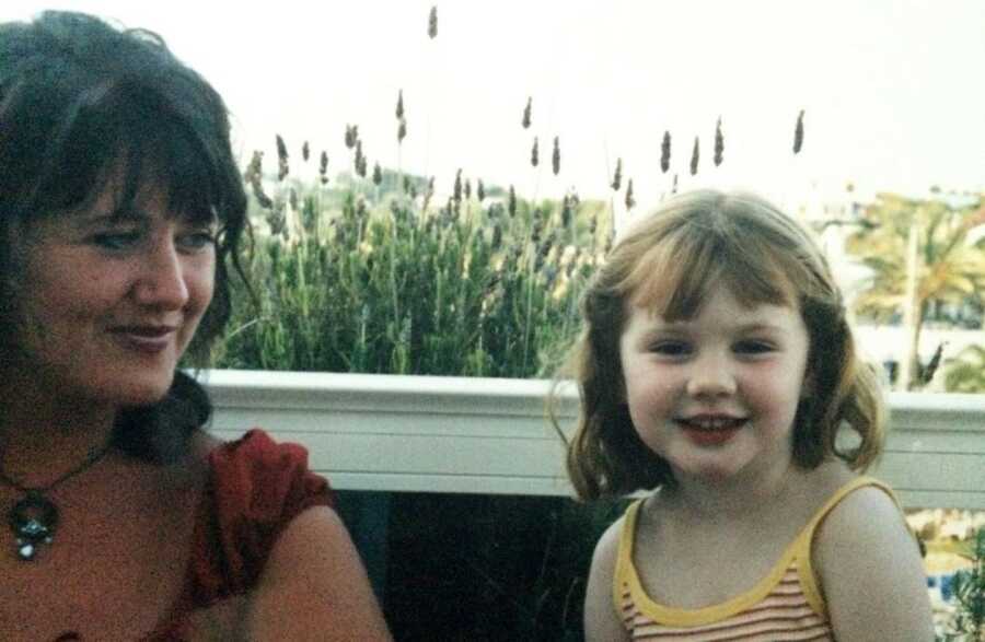 a dark-haired mom and her young daughter sit by a lake in front of plants