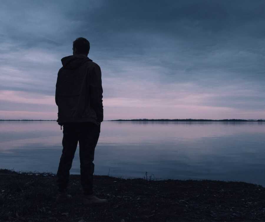 The silhouette of a man appears against a dark sky reflecting on a lake.