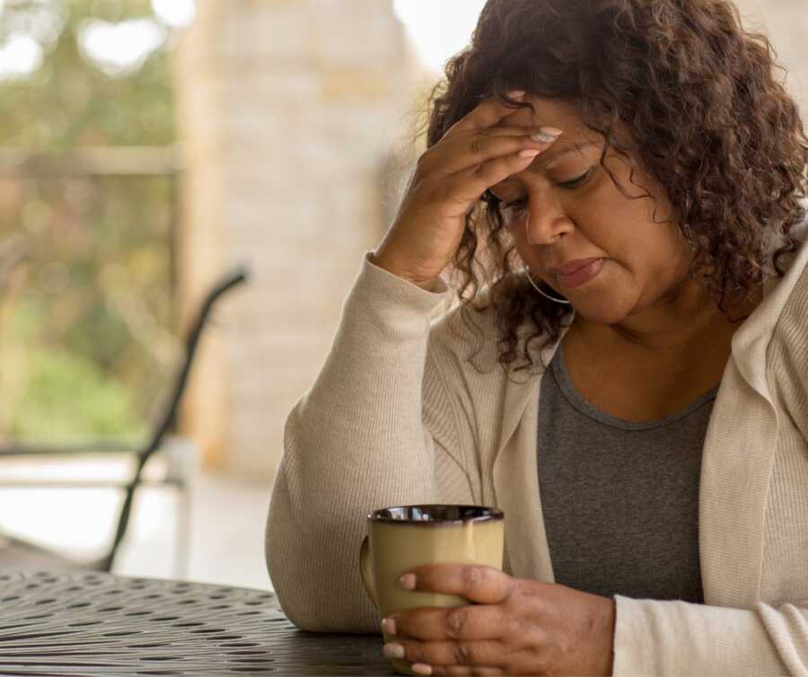 Tired, frustrated mom sits outside with coffee cup and hand on her forehead. 