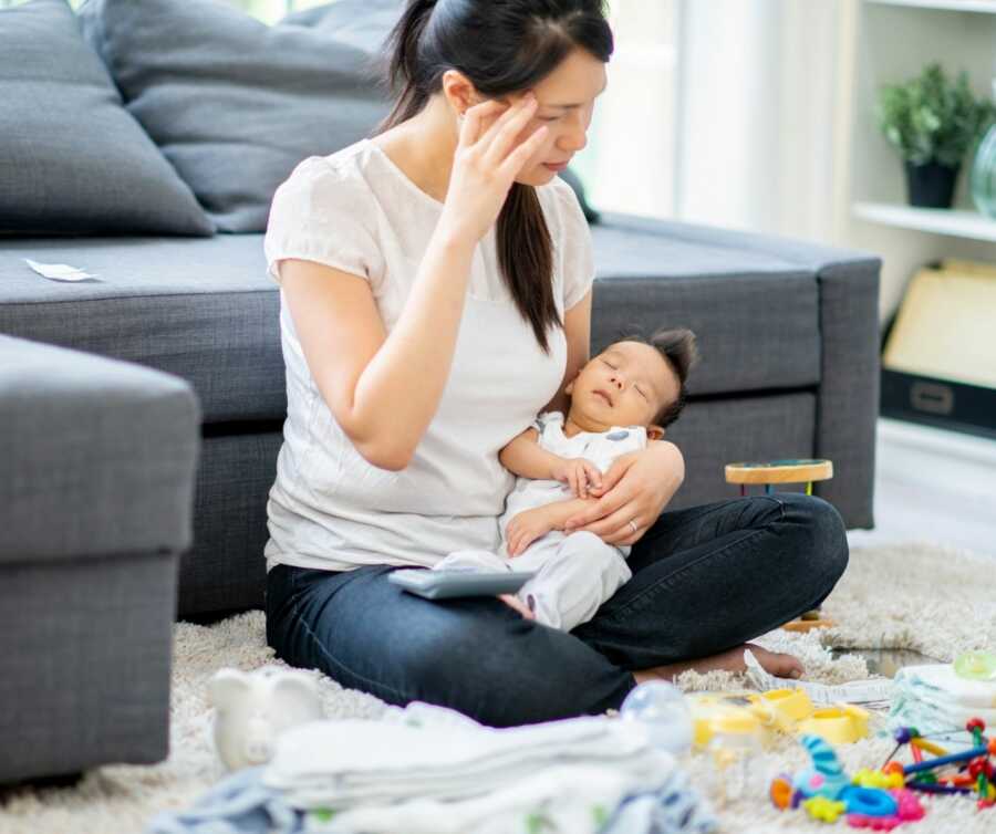 Young mom wipes tears from eyes while holding newborn baby.