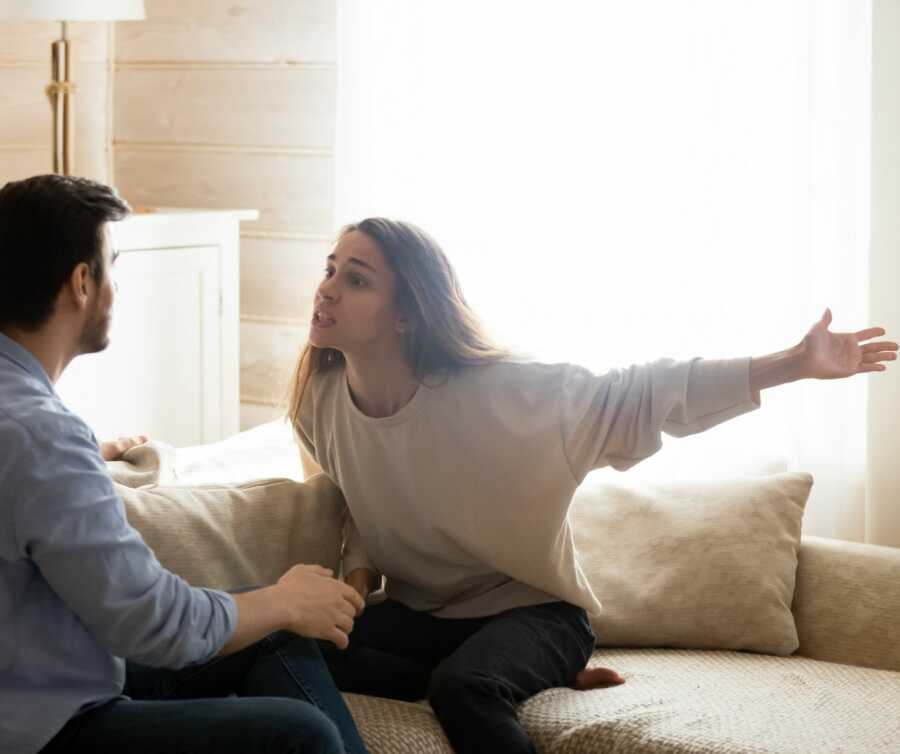 Young woman angrily yells and gestures at husband sitting on the couch.