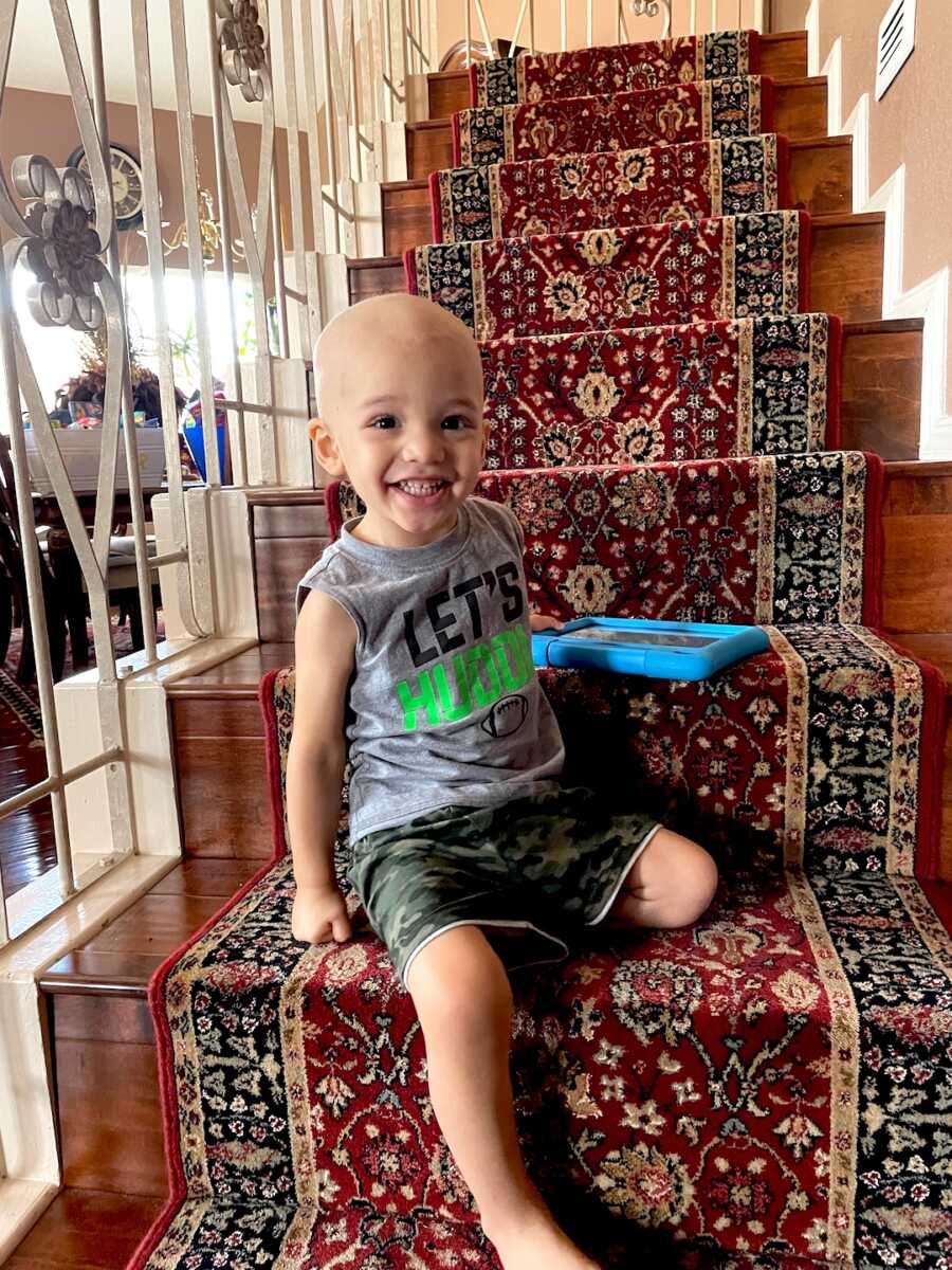 boy with childhood cancer sits on the stairs with his iPad and a smile