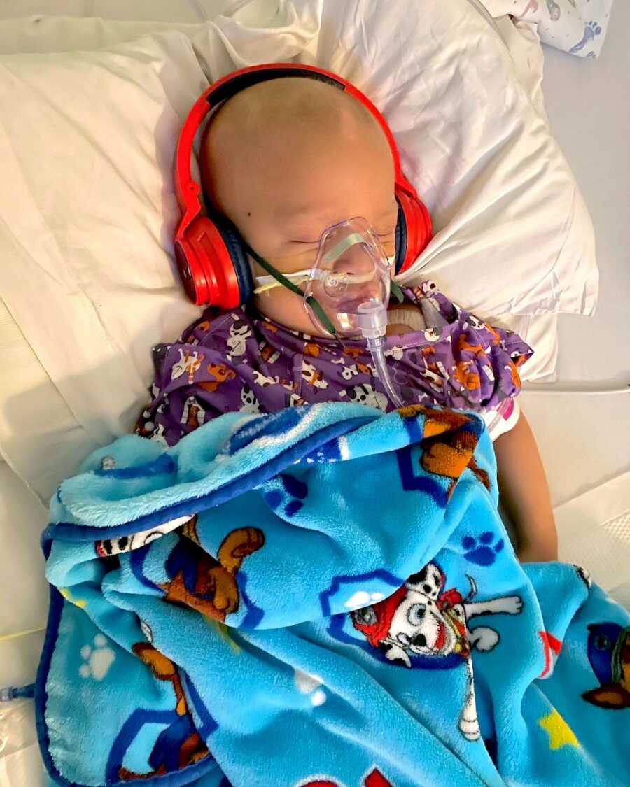 boy with childhood cancer sits in the hospital for treatments with an oxygen mask and headphones