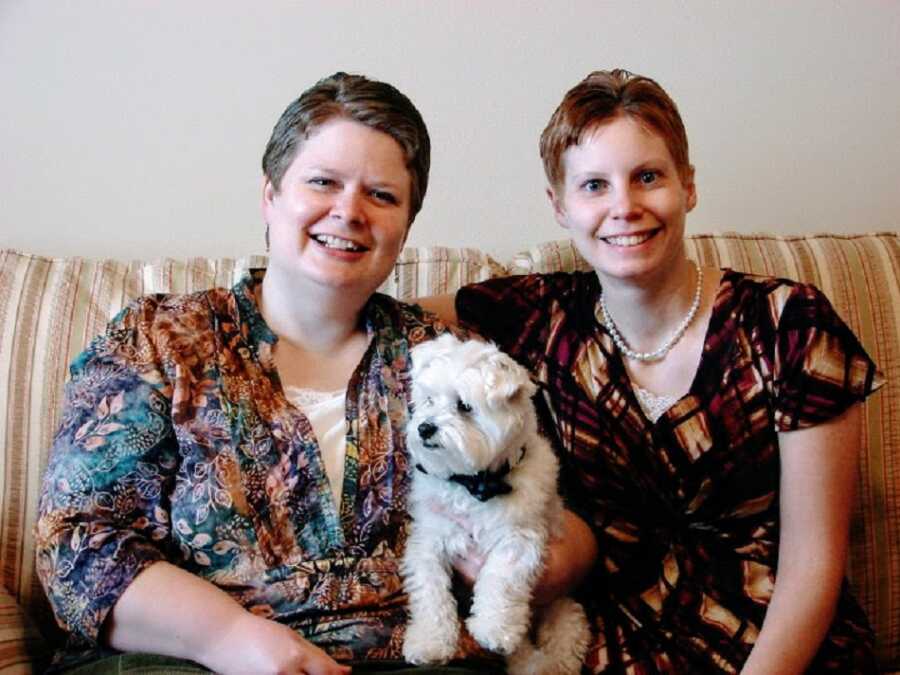 A lesbian couple sit together on the couch with a small white dog