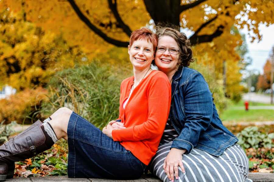 A couple sitting on a park bench in front of fall foliage at their engagement