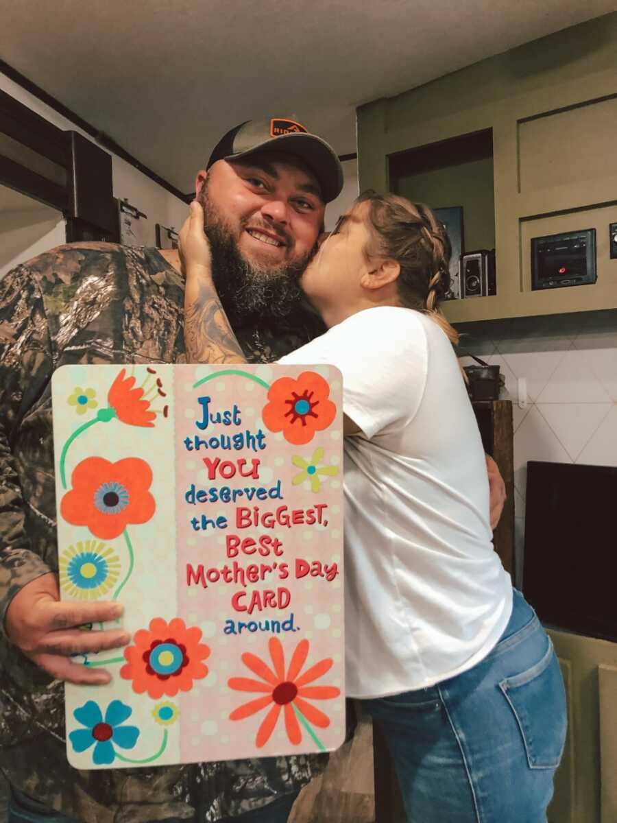 Wife kisses husband holding giant mother's day card for her.