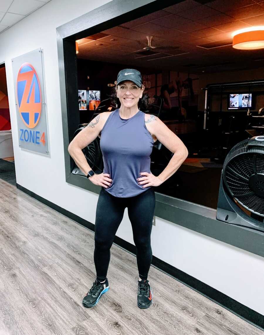 A woman stands in a gym wearing a tank top with her hands on her hips