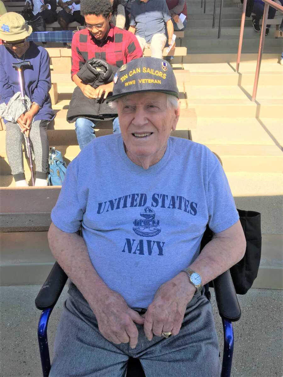 Old veteran wearing a blue U.S. Navy shirt and baseball cap sits in a chair in the shade. 