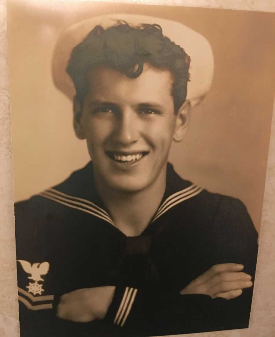 Young man in U.S. Navy sailor uniform crosses his arms and smiles in old photograph.