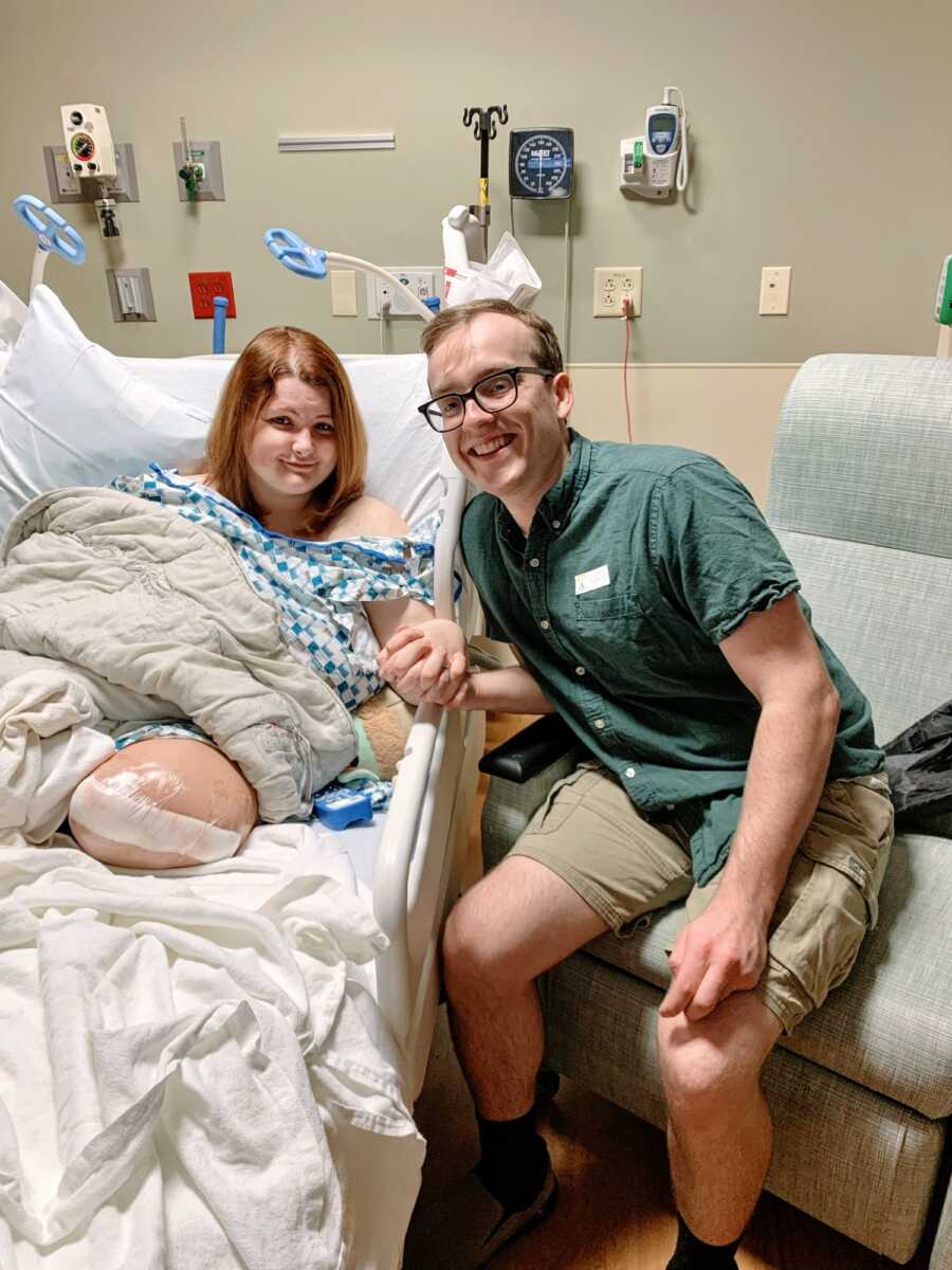 An amputee sits in a hospital bed holding her partner's hand