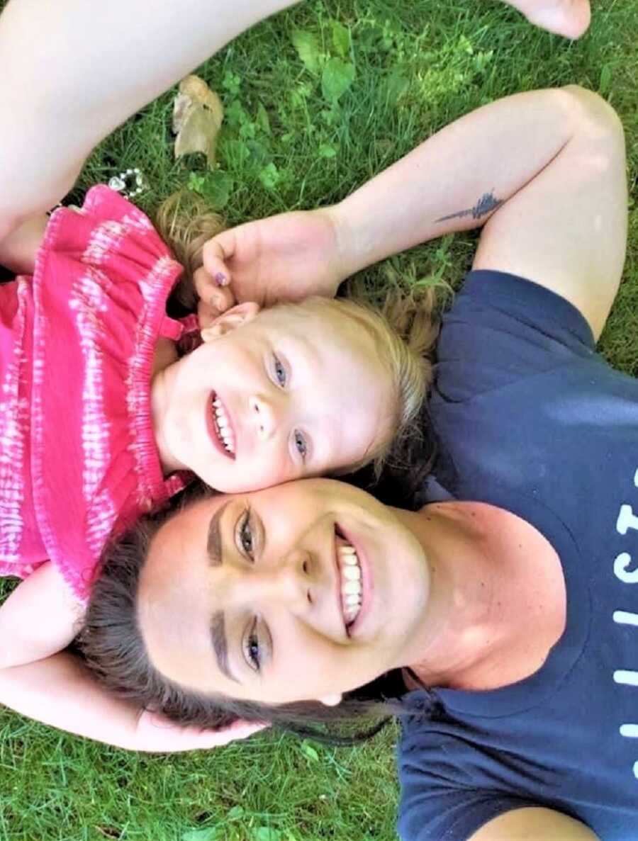 mom and toddler daughter laying on the grass with their head together and smiling 