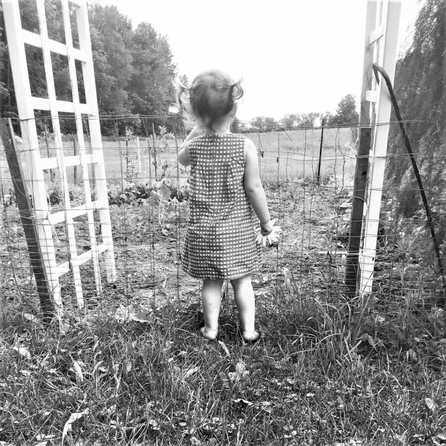 black and white image of toddler girl standing in front of a wire fence looking at a crops while holding a flower on her hand