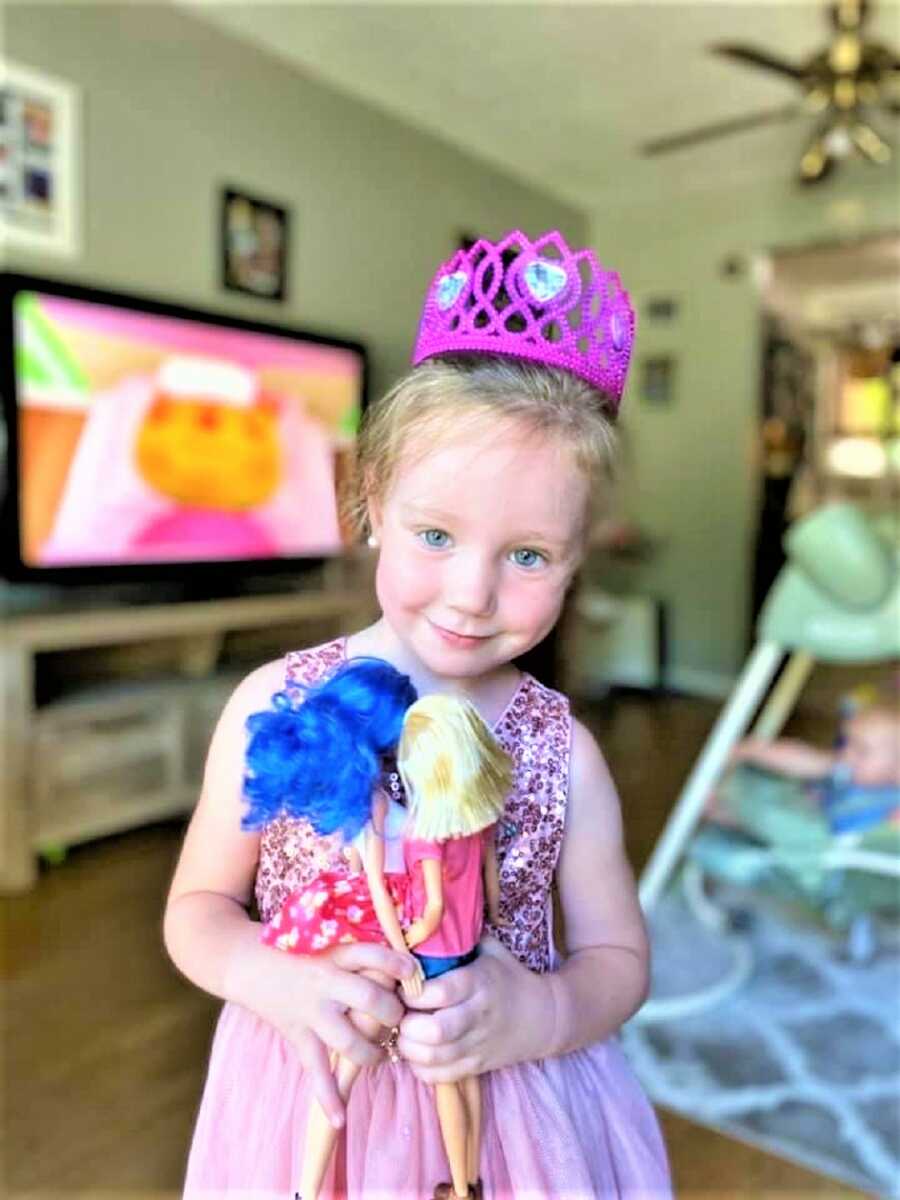 Toddler girl wearing a pink princess costume with a pink tiara playing with dolls in the Livingroom 