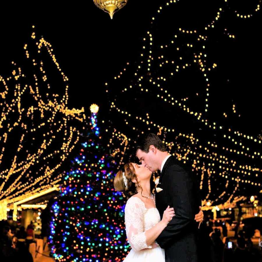 Husband and wife kissing on their wedding day with trees with Christmas lights on in the background