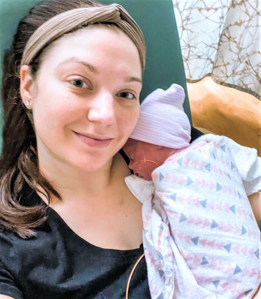 Selfie of mom holding one of her newborn twin girls on her shoulder while sitting on a chair at the NICU 