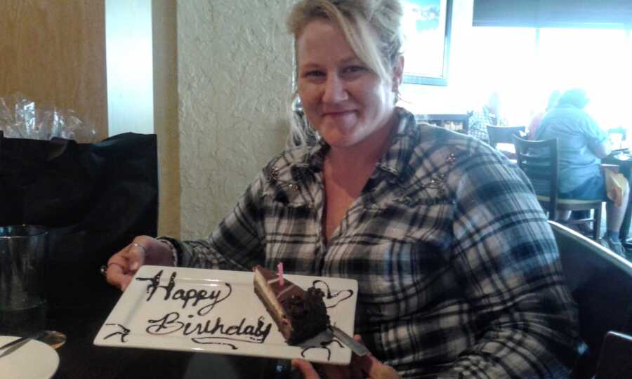 Woman in plaid shirt holds up slice of cake with "Happy birthday" written on the plate in chocolate sauce