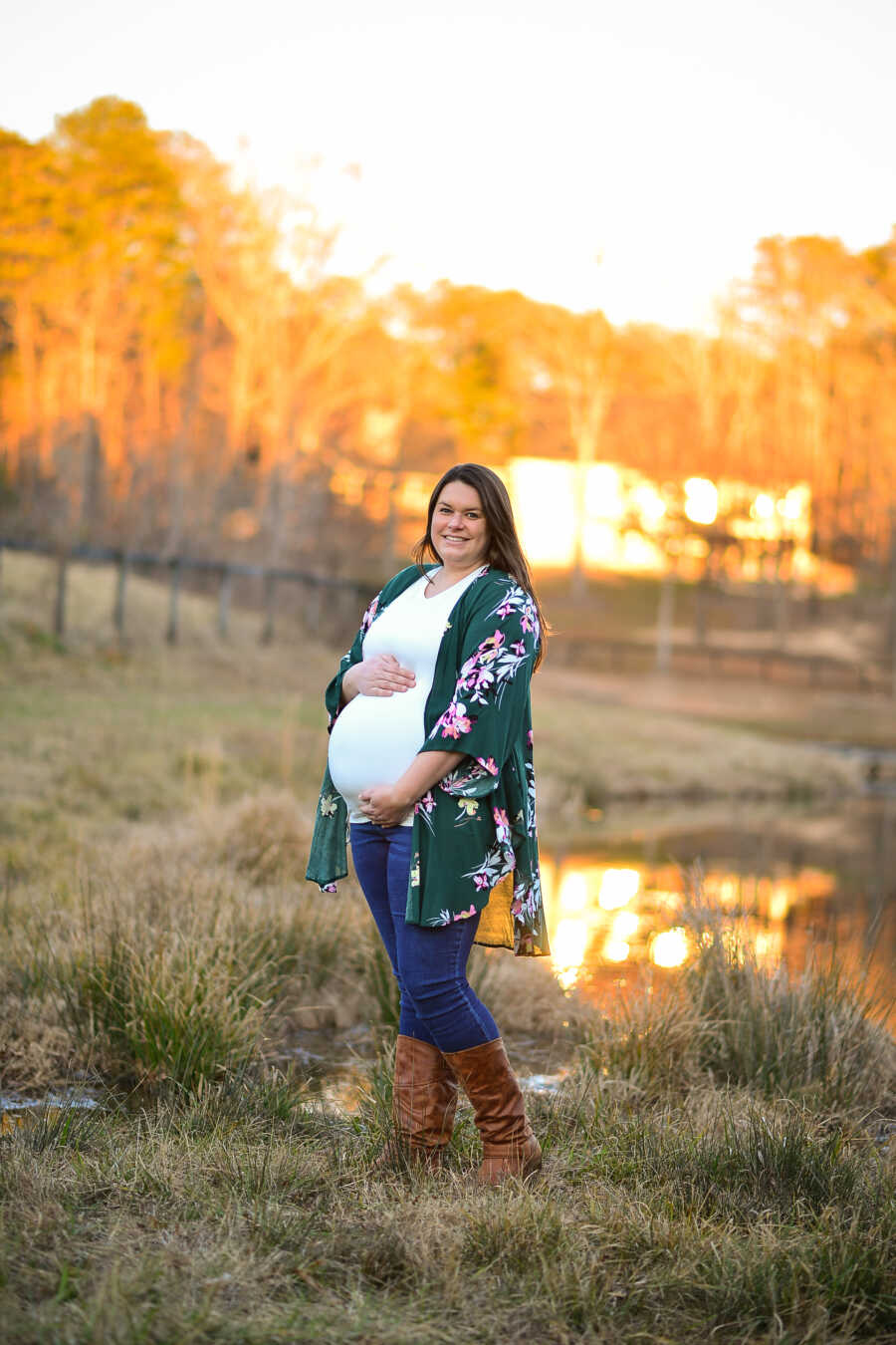 professional photo of a pregnant woman wearing a flower print cardigan and boots holding her baby bump with the sun setting in the back 