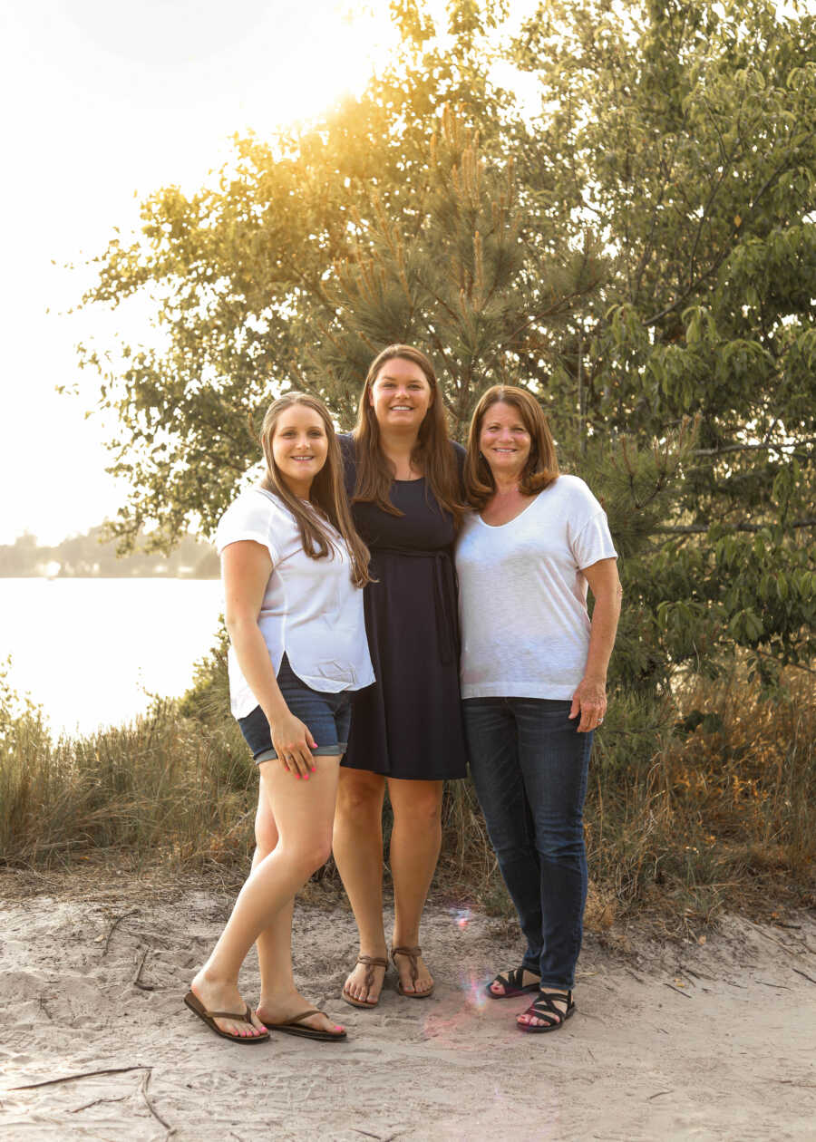 outdoors portrait of a mom and her two adult daughters standing together in front of a lake with trees in the back