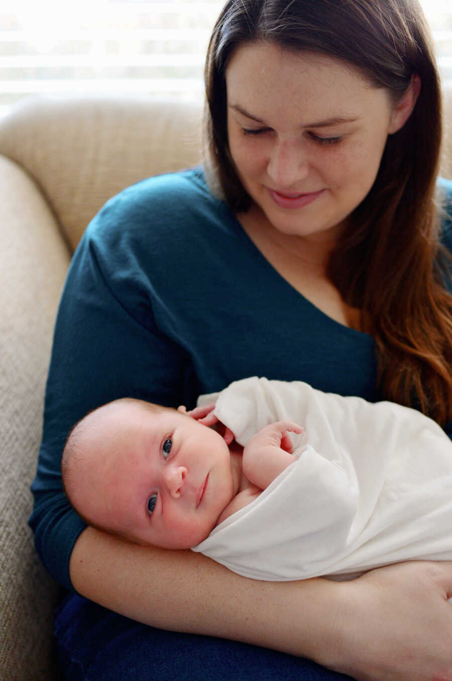 new mom sitting on a couch looking down at her newborn baby boy who is laying on her arms 