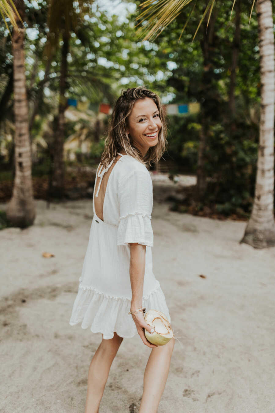 woman walking in the beach with half a coconut in her hand wearing a short white dress