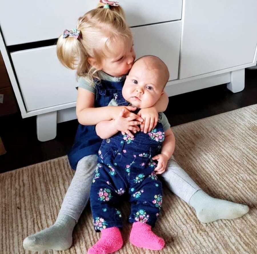 Big sister holds her little sister while giving her a kiss on the forehead