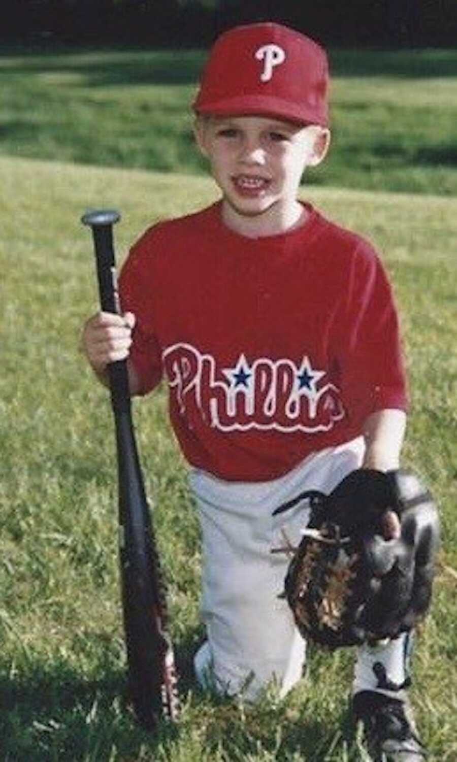 Young boy holding a baseball bat and glove. Wearing red shirt, kneeling in the grass