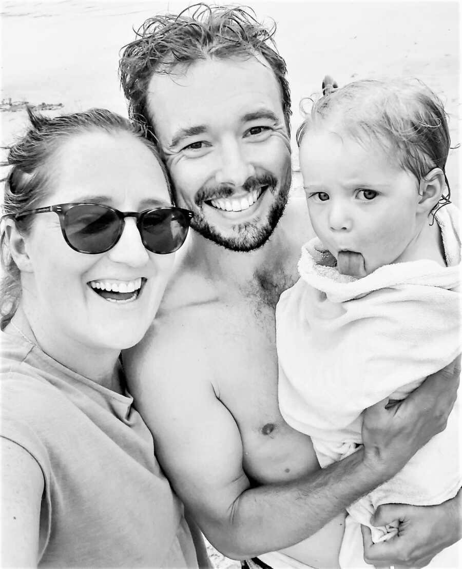 black and white selfie of mom and dad at the beach carrying their toddler daughter in a towel and smiling