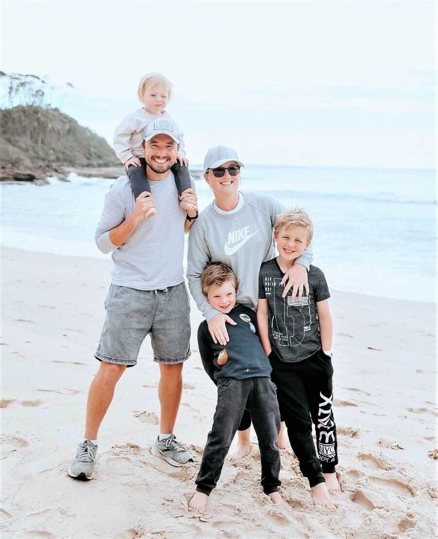 Portrait of family at the beach with mom resting her hands around her young sons' shoulders and dad carrying their toddler daughter on top of his shoulders