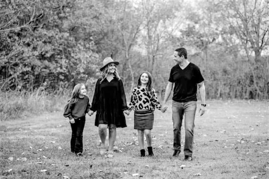 Black and white portrait of a mom, dad and their two daughter walking through a forest holding each others' hands and smiling 