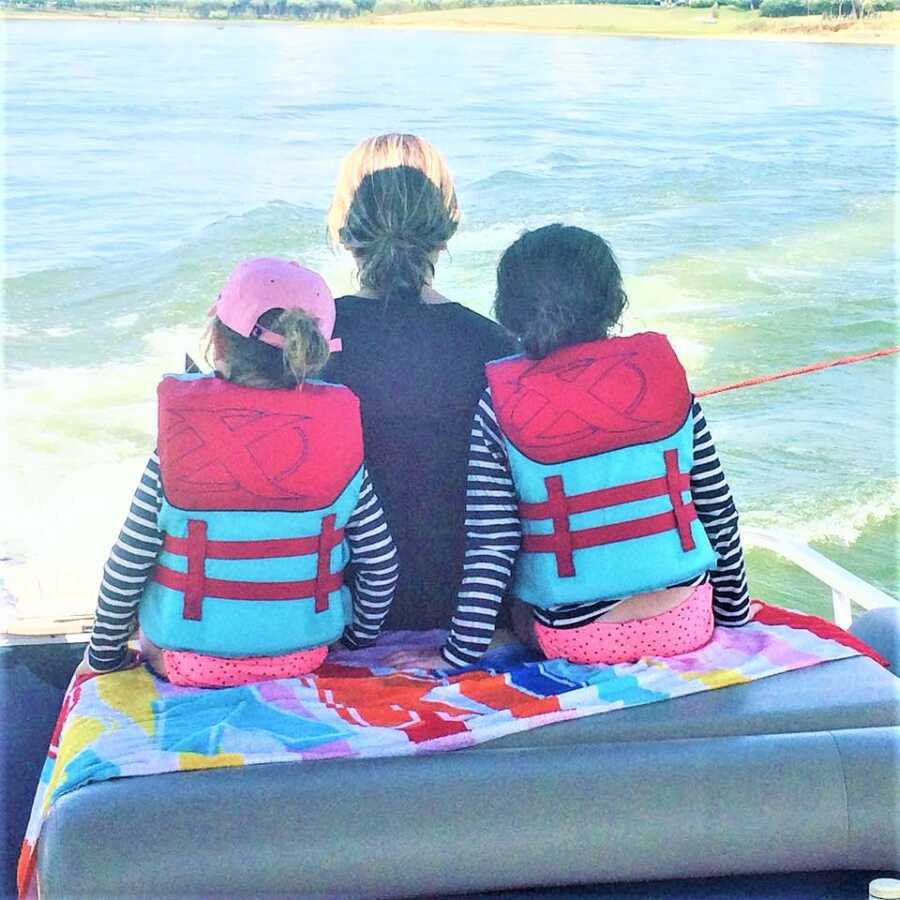 Picture taken from behind of a mom sitting on a boat with her two daughters facing the ocean 