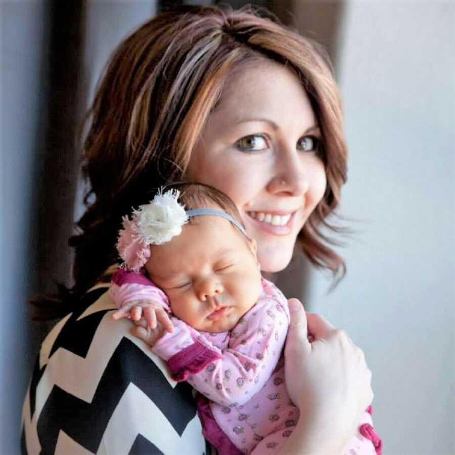 close-up of a mom holding her newborn daughter on her shoulder while looking at the camera smiling 