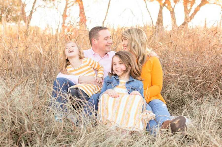 family portrait of mom, dad, and their two daughters sitting on the grass with the sun shining in the back while they all laugh 