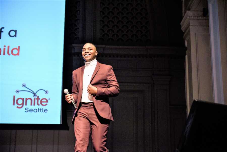 Black man wearing a brown suit giving a public speech in a stage 